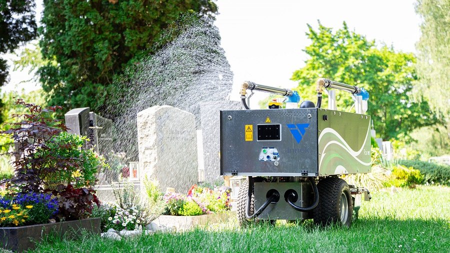 Auf dem Bild ist ein Roboter mit vier Rädern vor einer Grabanlage zu sehen. Der Roboter gießt das Grab.