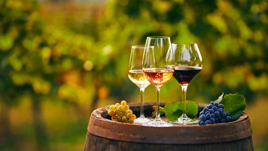 Drei edle Weingläser stehen auf einem Weinfaß. Ein Glas voller Rotwein, Weißwein und Rosé. Im Hintergrund sind Reben.